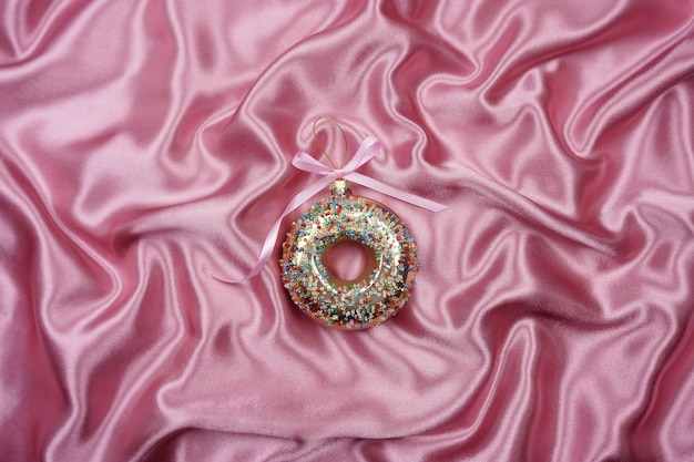 On a pink silk background there is a christmas tree toy in the form of a sweet donut