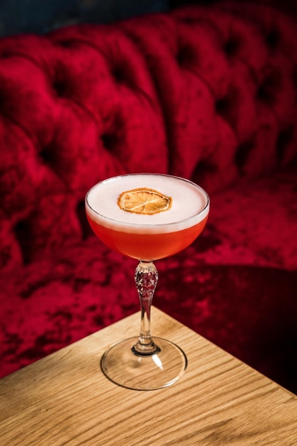 A pink sour alcoholic cocktail with foam garnished with dried lemon wheel served in a coupe glass on a wooden table of restaurant or bar vertical photo