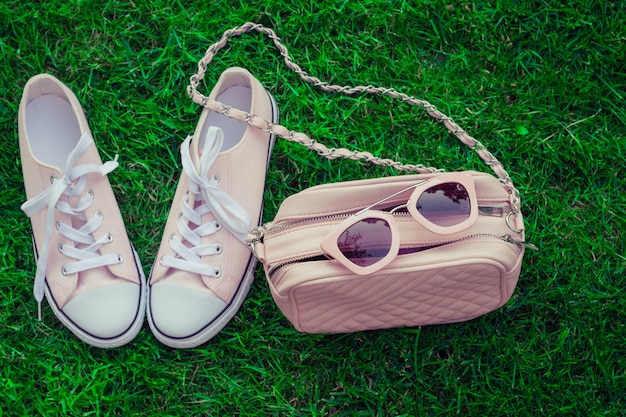 Pink sunglasses on a pink purse and sneakers with green grass as background