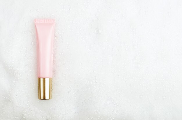 Pink tube with gel or face cream with a golden cap on a background of white foam with bubbles. Copy space, top view, flat lay.