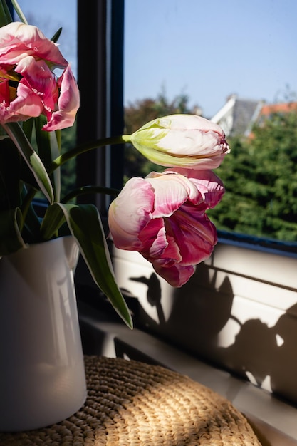 Pink tulip flower bouquet with green fresh stems in a white porcelain jar vase on a white window frame with blue sky background Vibrant colourful botany floral home decor idea