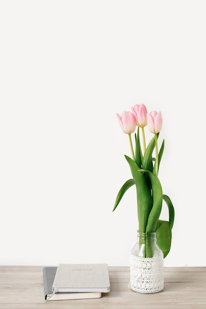 Photo pink tulip flowers in a vase on the table and a diary with a notepad, a white wall as a background with copying space