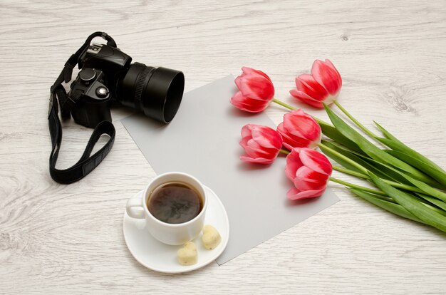 Pink tulips on a blank sheet of paper, mug of tea and a camera, a light wooden background.