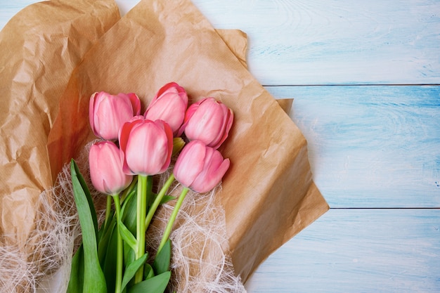 Розовые тюльпаны лежат на крафтовой бумаге на синем деревянном фоне. Плоская планировка, вид сверху