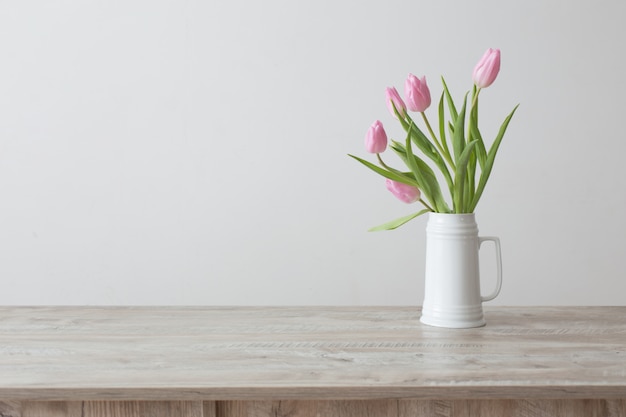 Pink tulips in white ceramic jug on wooden table on background white wall