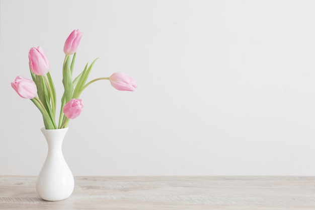 Pink tulips in white ceramic vase on wooden table on background white wall