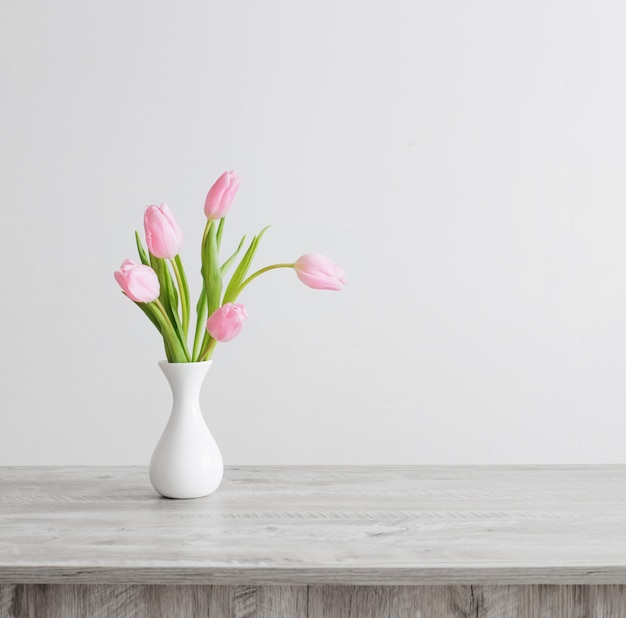 Pink tulips in white ceramic vase on wooden table on background white wall