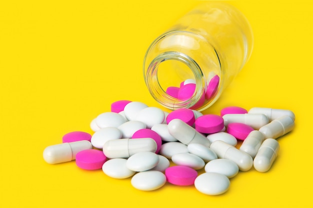 Pink and white tablets scattered from a glass jar on a yellow background