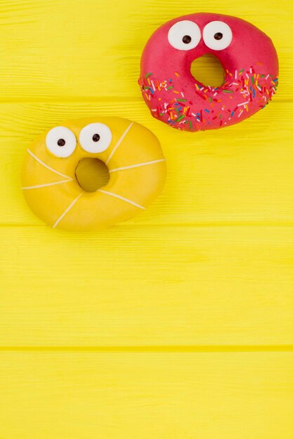 Pink and yellow donuts, top view. Donuts with surprised eyes on color wooden background. Space for text.