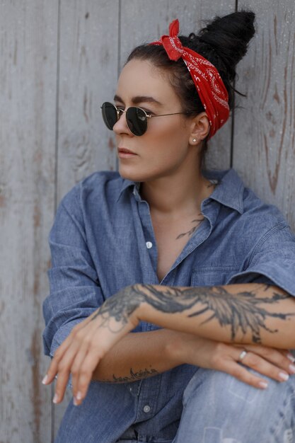 Pinup style. Stylish woman with a bandana and sunglasses in fashion jeans clothes sits near a wooden wall. Tattoo on the hand