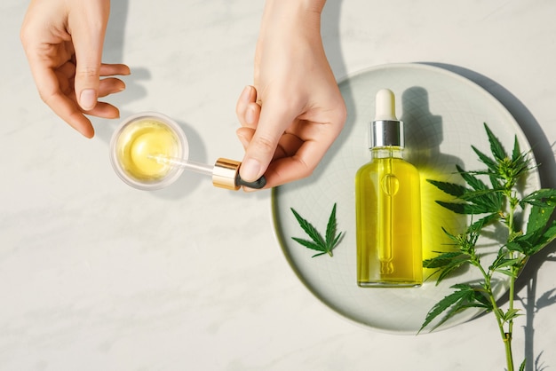 Pipetta con olio cosmetico cbd in mani femminili su un tavolo con una bottiglia di olio di cannabis e foglie di canapa, marijuana