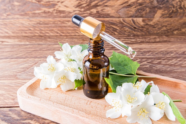 A pipette with natural jasmine oil lies on an open bottle\
standing on a brown wooden tray among the flowers of jasmine\
natural cosmetics