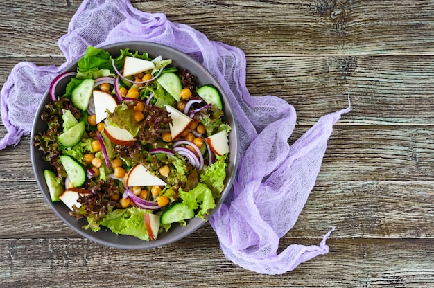 Foto pittige gezonde salade van verse groenten, rode ui, kikkererwten, gerookte kaas (worst kaas), komkommers in een kom op een houten tafel. vegetarisch gerecht. bovenaanzicht.