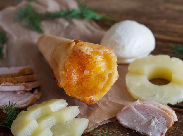 The pizza cone lies on paper on a wooden table. This is a pineapple, chicken and mushroom pizza. Street food