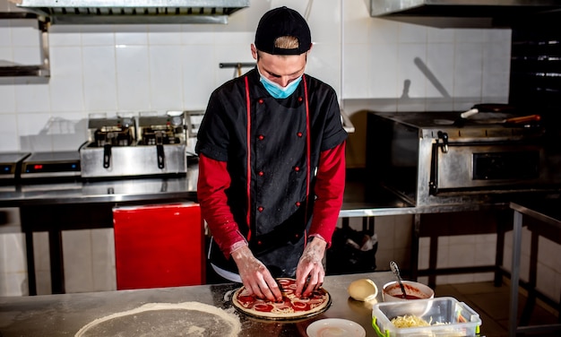 Pizza cooking concept, chef in medical mask puts ingredients on pizza