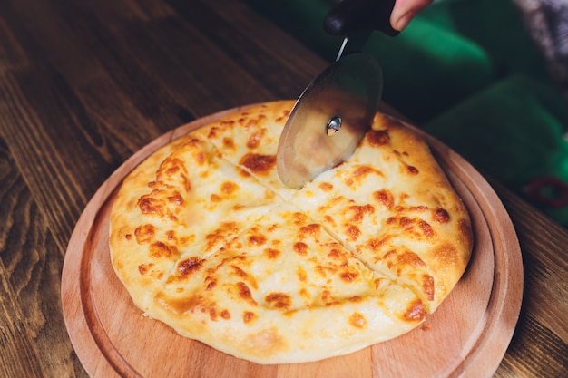 Pizza cutter wheel slicing ham pizza with capsicum and olives on wooden board on table.