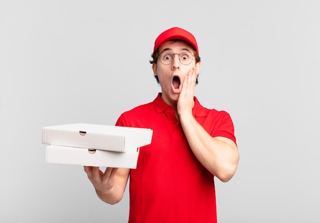 Pizza deliver boy feeling shocked and scared, looking terrified with open mouth and hands on cheeks