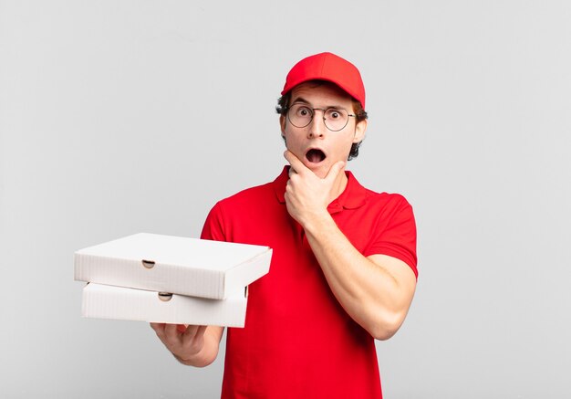 Pizza deliver boy with mouth and eyes wide open and hand on chin, feeling unpleasantly shocked, saying what or wow