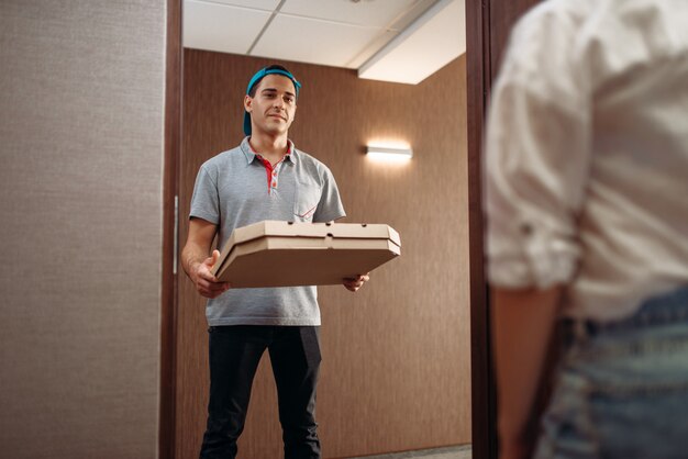 Ragazzo delle consegne di pizza con scatola di cartone in mano sulla soglia di casa, offrendo servizio. corriere da pizzeria e cliente al chiuso