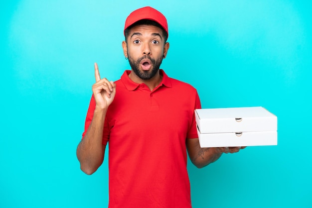 Consegna pizza uomo brasiliano con uniforme da lavoro che raccoglie scatole di pizza isolate su sfondo blu con l'intenzione di realizzare la soluzione sollevando un dito