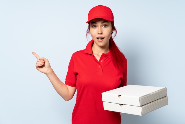 Pizza delivery girl holding a pizza over isolated background surprised and pointing finger to the side