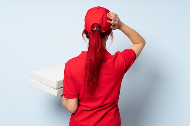 Pizza delivery girl holding a pizza over isolated wall in back position and thinking