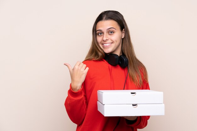 Pizza delivery girl holding a pizza over isolated wall pointing to the side to present a product