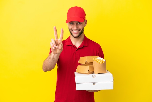 Pizza delivery man picking up pizza boxes and burgers over isolated background smiling and showing victory sign