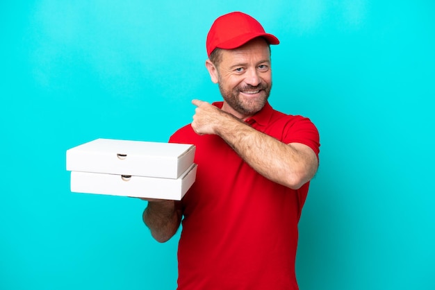 Pizza delivery man with work uniform picking up pizza boxes isolated on blue background pointing back