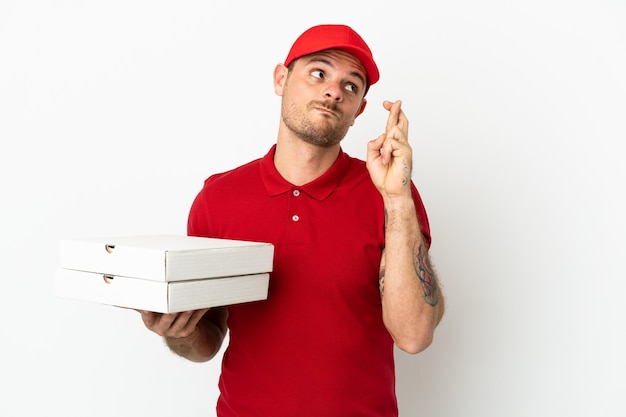 Pizza delivery man with work uniform picking up pizza boxes over isolated  white wall with fingers crossing and wishing the best