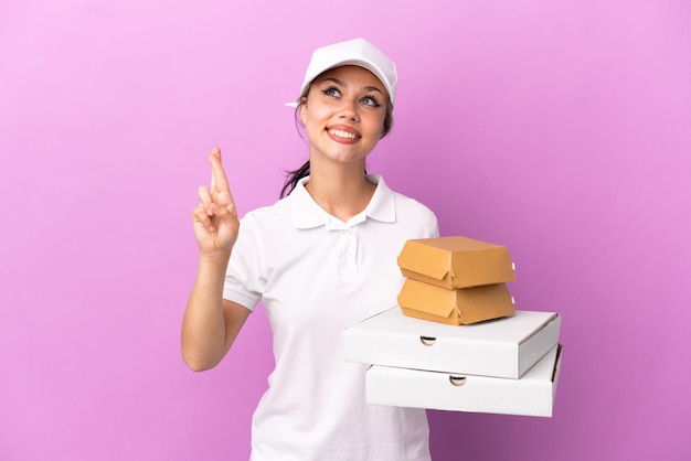 피자 배달 러시아 소녀는 작업복을 입고 보라색 배경에 격리된 피자 상자와 햄버거를 줍고 손가락을 교차하고 최고를 기원합니다.