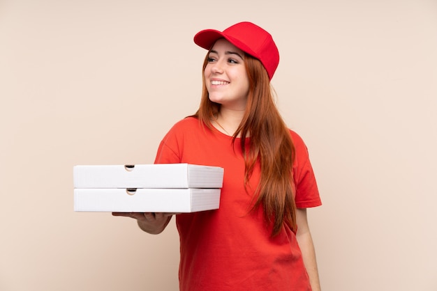 Pizza delivery teenager girl holding a pizza over isolated wall laughing