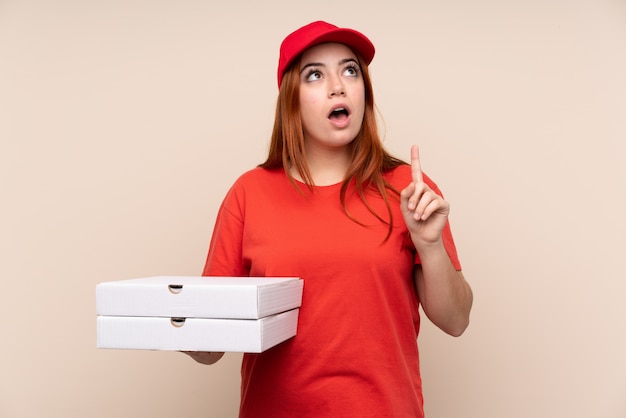 Pizza delivery teenager woman holding a pizza over isolated wall pointing with the index finger a great idea