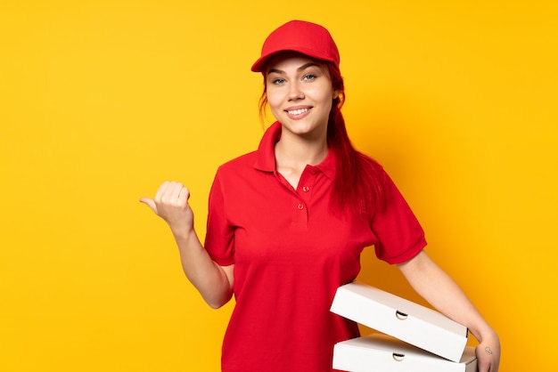 Pizza delivery woman holding a pizza over isolated wall pointing to the side to present a product