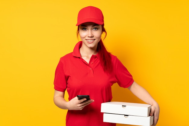 Pizza delivery woman holding a pizza over isolated wall sending a message with the mobile