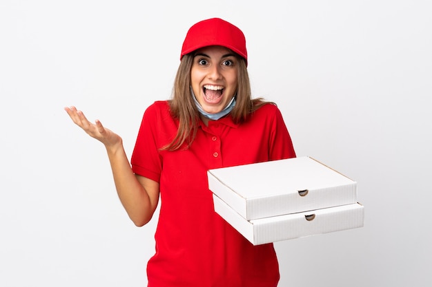 Pizza delivery woman holding a pizza and protecting from the coronavirus with a mask on isolated white wall with shocked facial expression