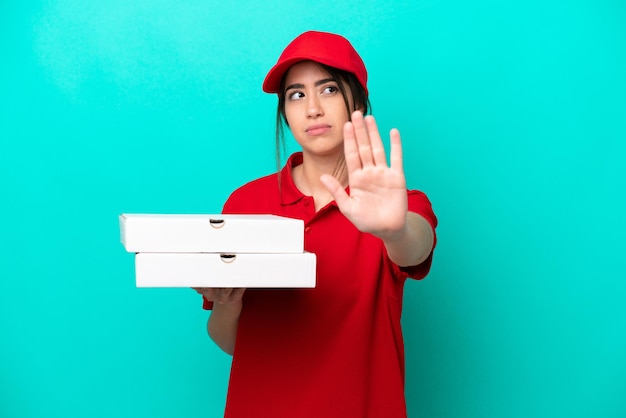 青い背景に分離されたピザの箱を拾う仕事の制服を着たピザ配達の女性は、停止ジェスチャーを作成し、失望しました