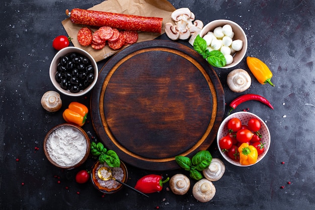 Pizza ingredients on the dark background and round cutting board. Pepperoni, mozzarella, tomatoes, olives, mushrooms and flour are different products for making pizza and pasta