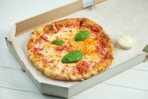 Pizza margherita with tomatoes, basil and mozzarella in a cardboard box with sauce for sides on a wooden table