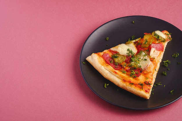 Pizza slice with pepperoni, salami, melted mozzarella cheese, pickles and dill in black plate on red background, angle view copy space