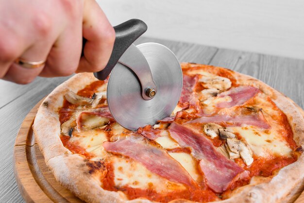 Foto pizza per affettare utilizzando il coltello a ruota pizza con prosciutto, salame, pomodori, insalata e parmigiano su una tavola di legno su un tavolo di vetro