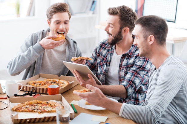 Pizza time! Three young cheerful men eating pizza together while sitting in the office