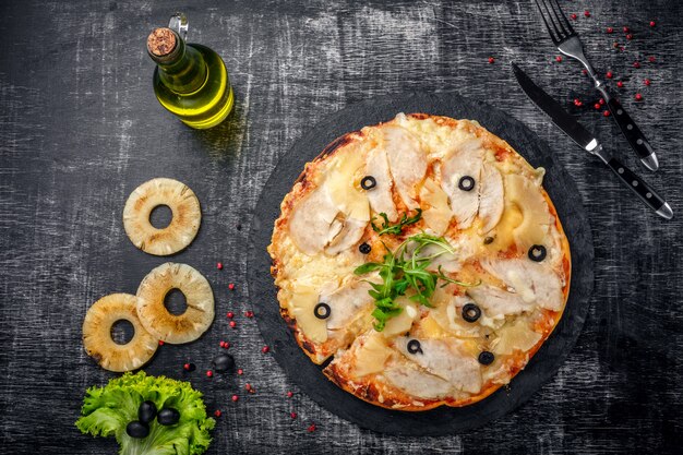 Pizza with chicken, pineapple, cheese and olives on stone and a black wooden scratched background