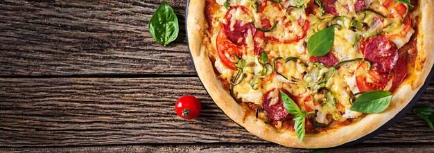 Pizza con pollo, salame, zucchine, pomodori ed erbe sul tavolo di legno d'epoca. . banner. cucina italiana