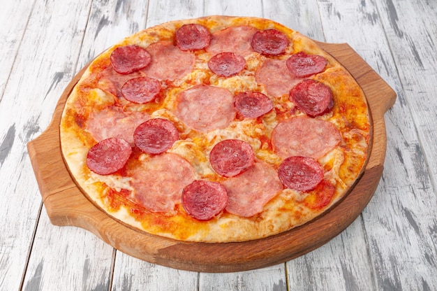 Pizza with ham, salami and mozzarella cheese. On a round wooden board. Light wooden background. View from above.