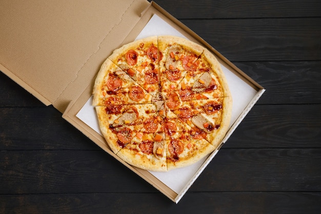 Pizza with pepperoni, ham, bell pepper and sesame on dark wooden table in cardboard package