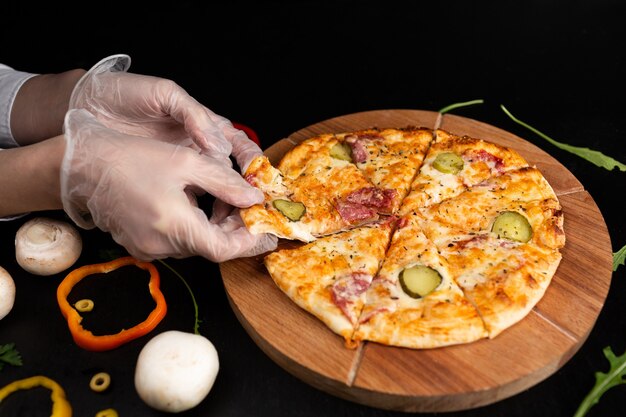 Pizza su un supporto di legno su una superficie nera le mani guantate prendono una fetta di pizza