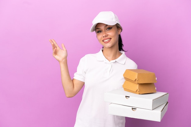 Pizzabezorger Russisch meisje met werkuniform oppikken van pizzadozen en hamburgers geïsoleerd op een paarse achtergrond die de handen naar de zijkant uitstrekt om uit te nodigen om te komen