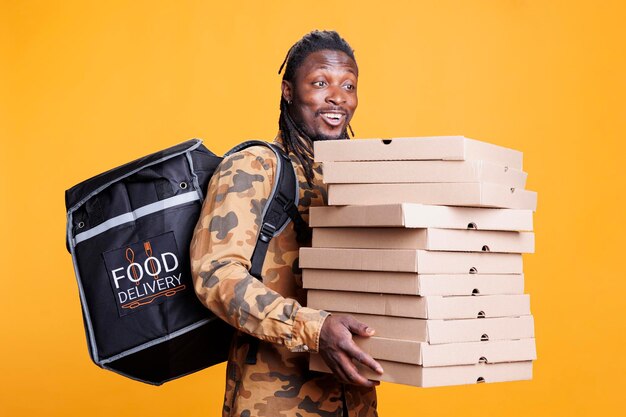 Pizzeria delivery employee holding cardboard stack full with pizza, delivering takeaway food order to client during lunch time. african american deliveryman carrying thermal backpack in studio