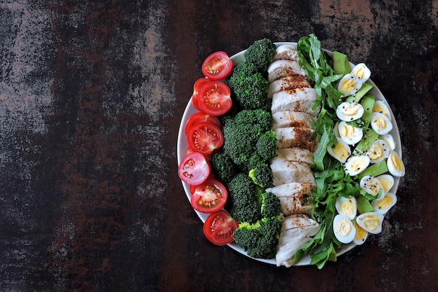 Plaat met een Keto dieetvoeding. Een set van producten voor het ketogene dieet op een bord. Cherrytomaatjes, gekookte broccoli, gestoomde kipfilet, salade met rucola, avocado en kwarteleitjes. Keto lunch.